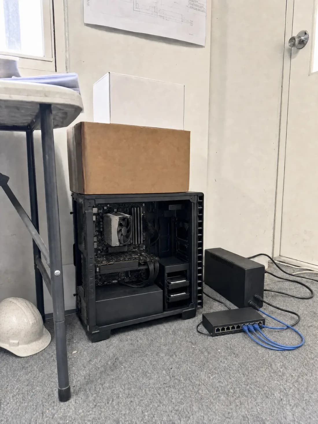 A construction.live server running on the floor of a site office, network switch and UPS beside it and a hard hat on the ground